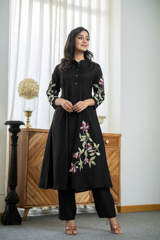 Woman wearing a black embroidered outfit standing in a room with a wooden cabinet and decorative items.